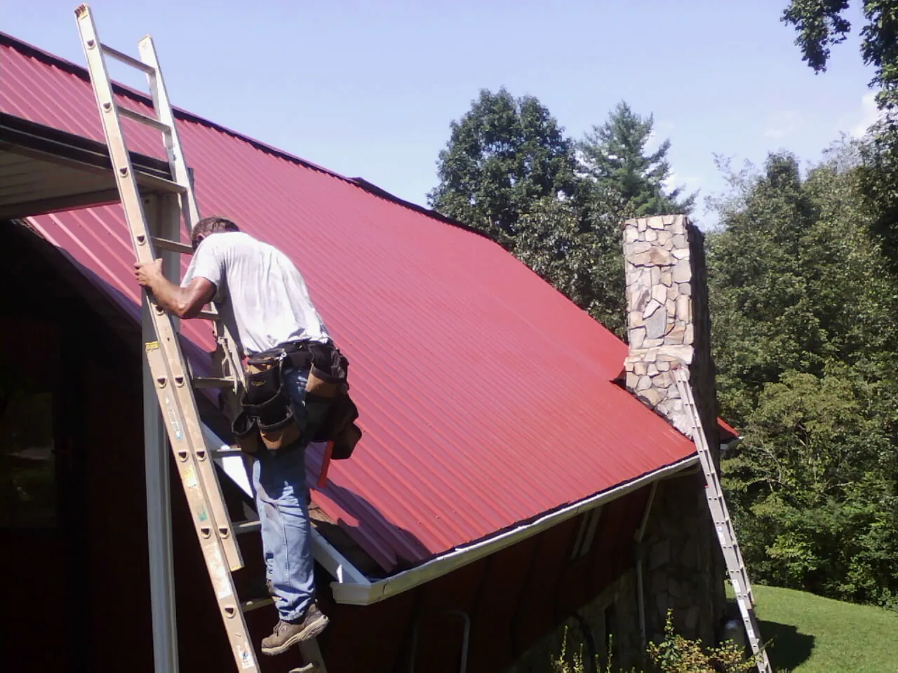 Roofer on a ladder beside a red metal roof and stone chimney — Soffit & Fascia Repair by Roofing Brothers in West Mifflin